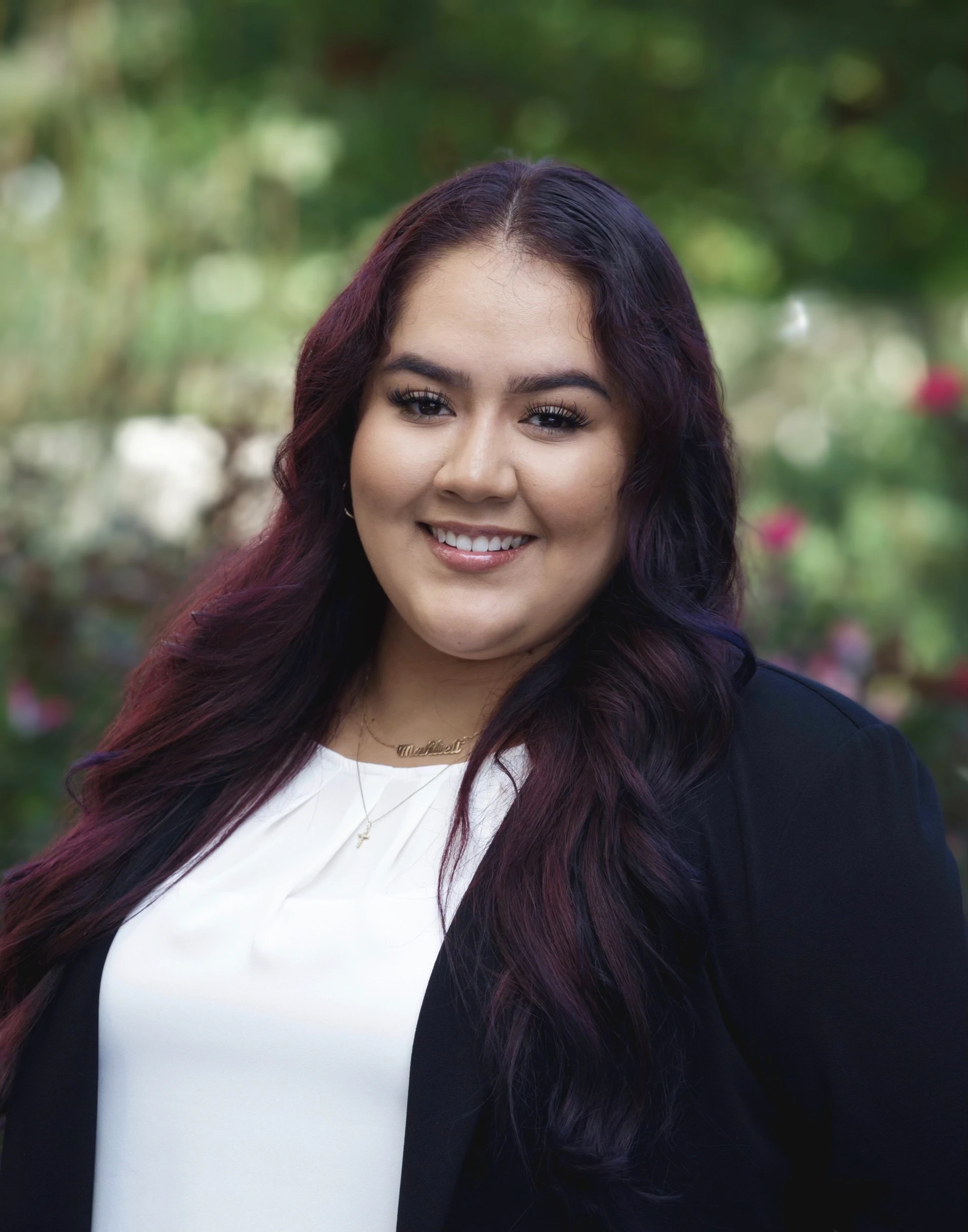 Uma pessoa com cabelo longo e escuro e uma expressão amigável está sorrindo, vestindo uma blusa branca e uma jaqueta preta, tendo ao fundo uma vegetação exuberante e flores coloridas desfocadas.