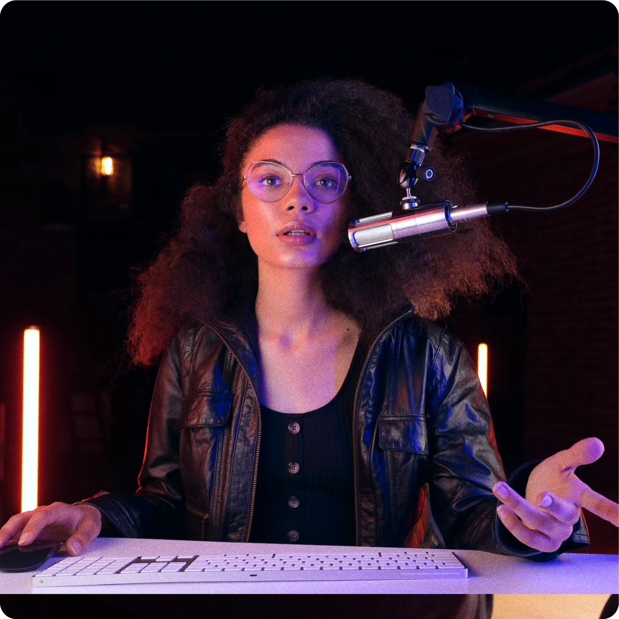 Image of a woman with curly hair and glasses, sitting at a desk with a keyboard and mouse. She is wearing a black leather jacket and is speaking into a professional microphone mounted on a boom arm. The background is dark with neon lights creating a vibrant, modern studio atmosphere, highlighting the woman's focused expression as she engages with her podcast or streaming audience.