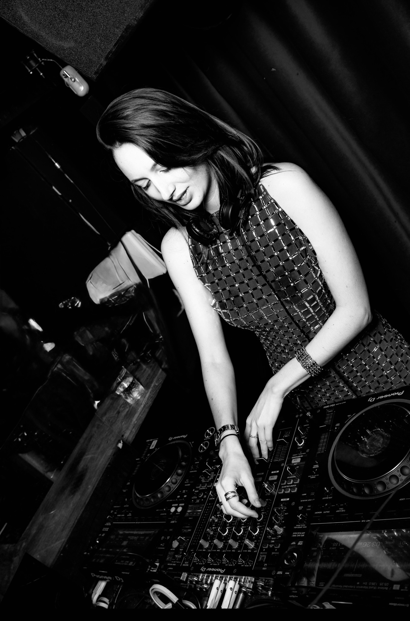 Photographie de Lizy en plein mix derrière ses platines, vêtue d'une robe texturée sans manches, dans une ambiance de club sombre.