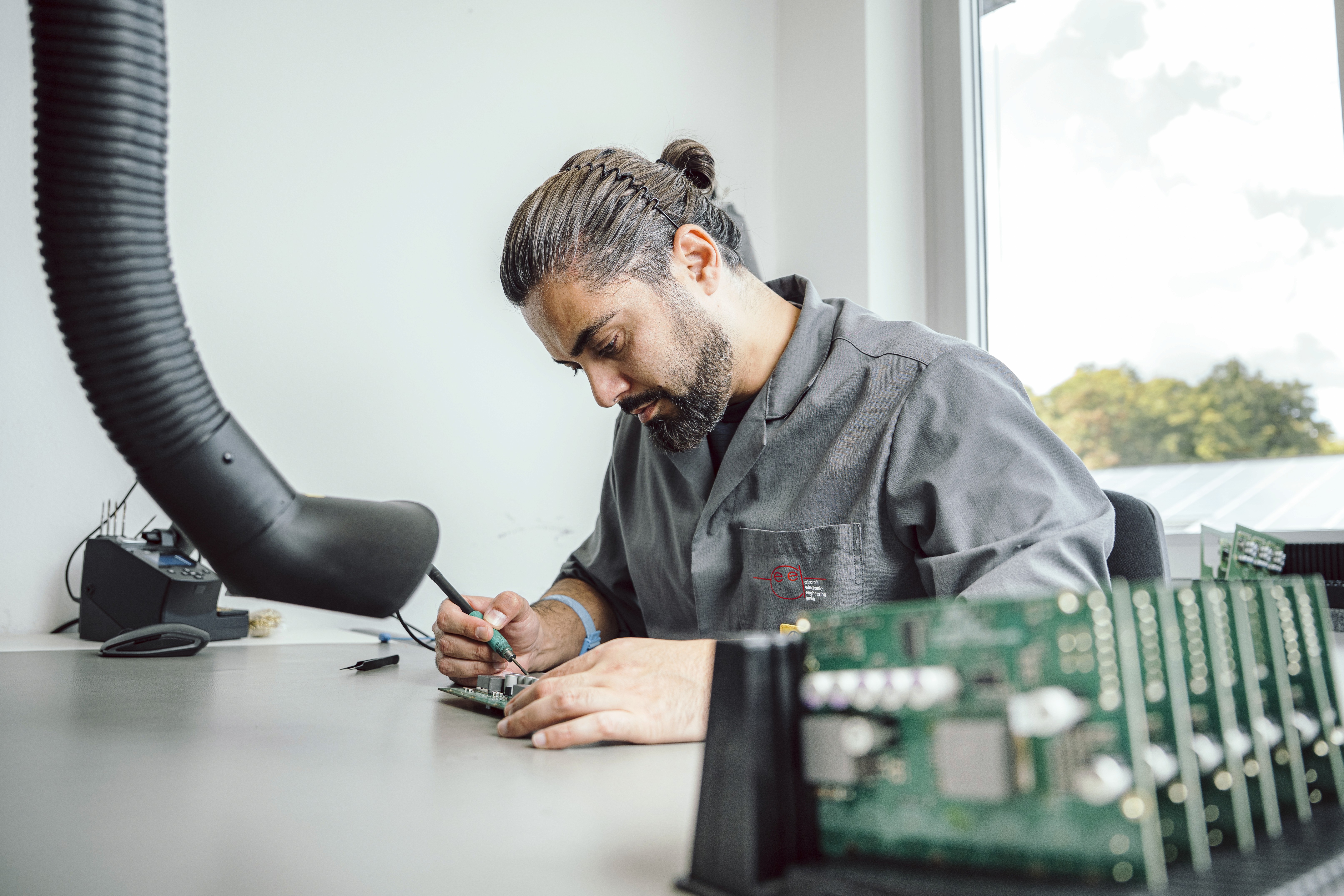 A man in work clothes solders a circuit board, with finished circuit boards visible and blurred in the foreground.