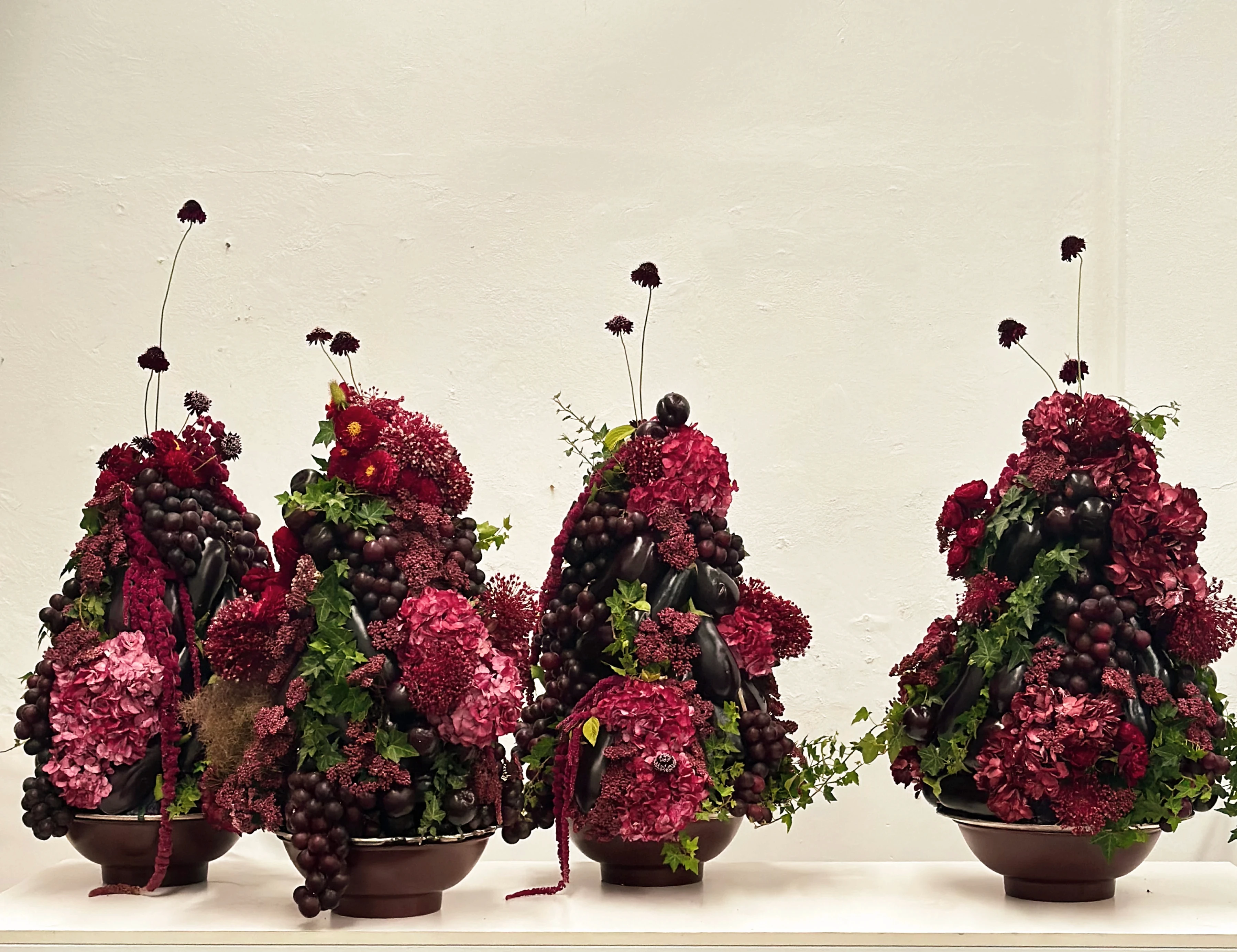 Cuatro centros de mesa abundantes con hortensias bordó, uvas y berenjenas sobre pedestales de madera.