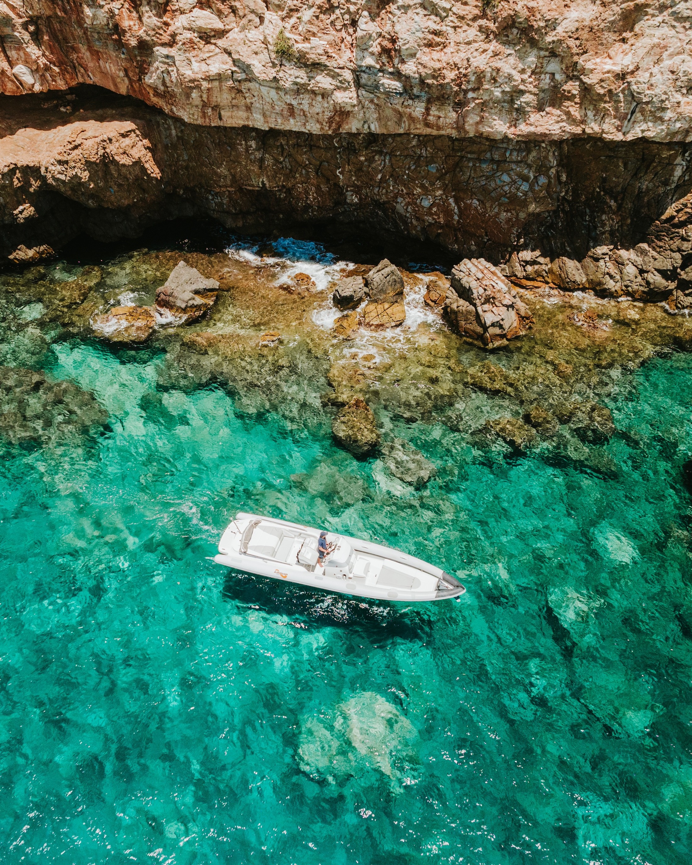 Aerial view of white motor yacht anchored in crystal-clear turquoise waters surrounded by dramatic limestone cliffs and rocky coastline.