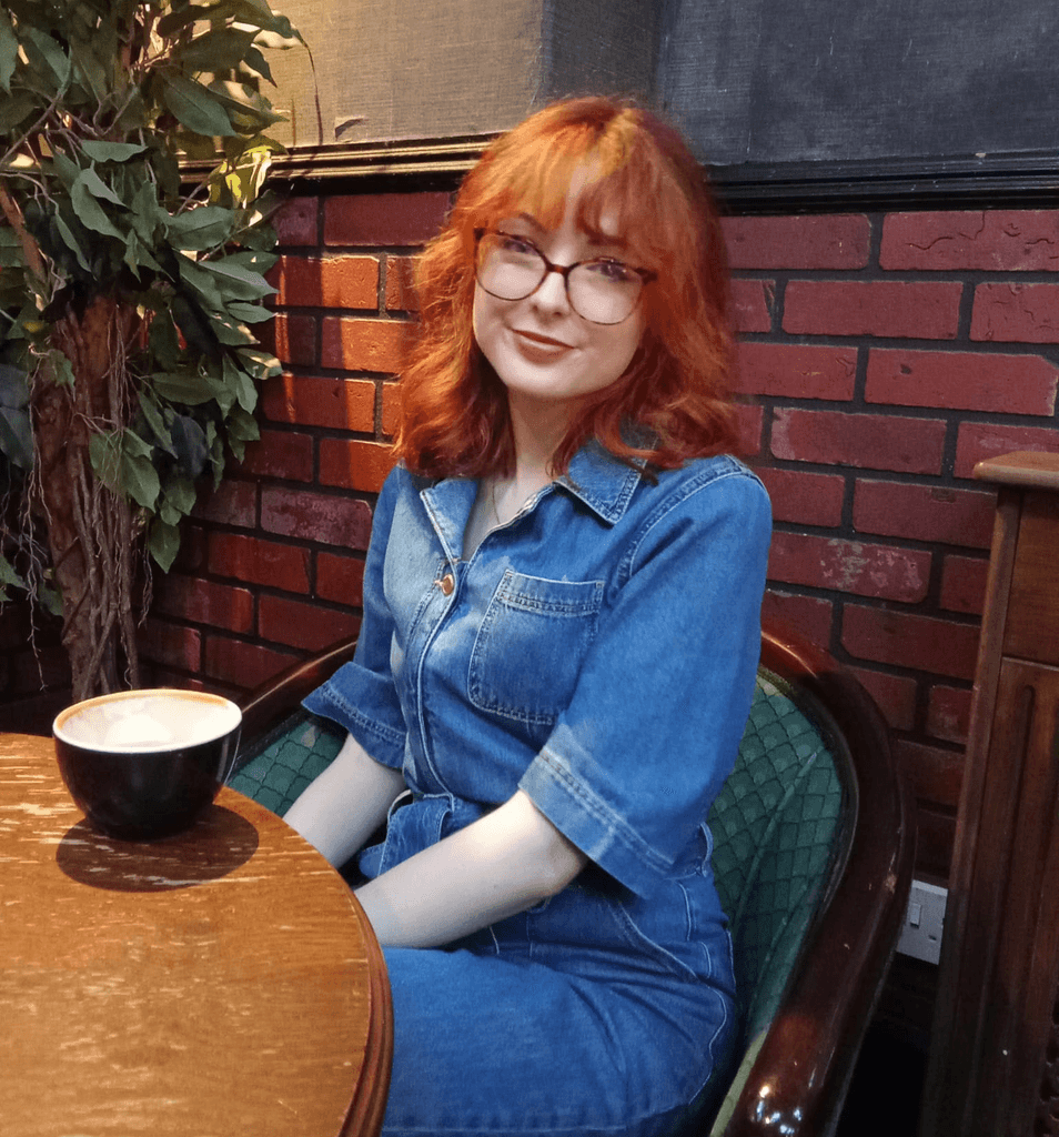 Amy sitting on a green chair with her back to a red bring wall, she is in a coffee shop and has a cappuccino on a wooden table in front of her.