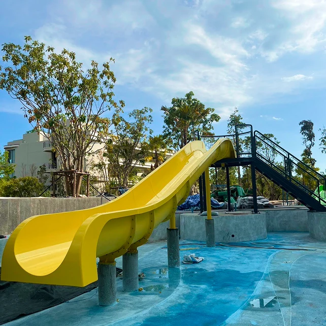 Một cầu trượt nước màu vàng tươi đổ vào khu vực hồ bơi trống, bao quanh bởi những cây xanh và bầu trời xanh trong vắt. Không khí rất yên bình và mời gọi.