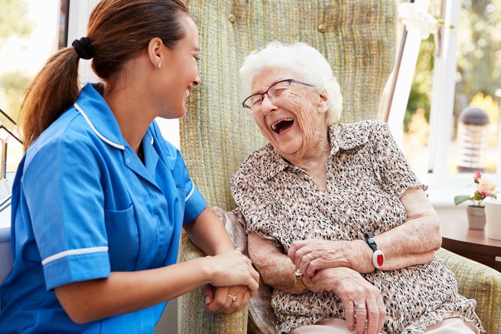 Caregiver and elderly woman sitting together, laughing and holding hands in a bright room.