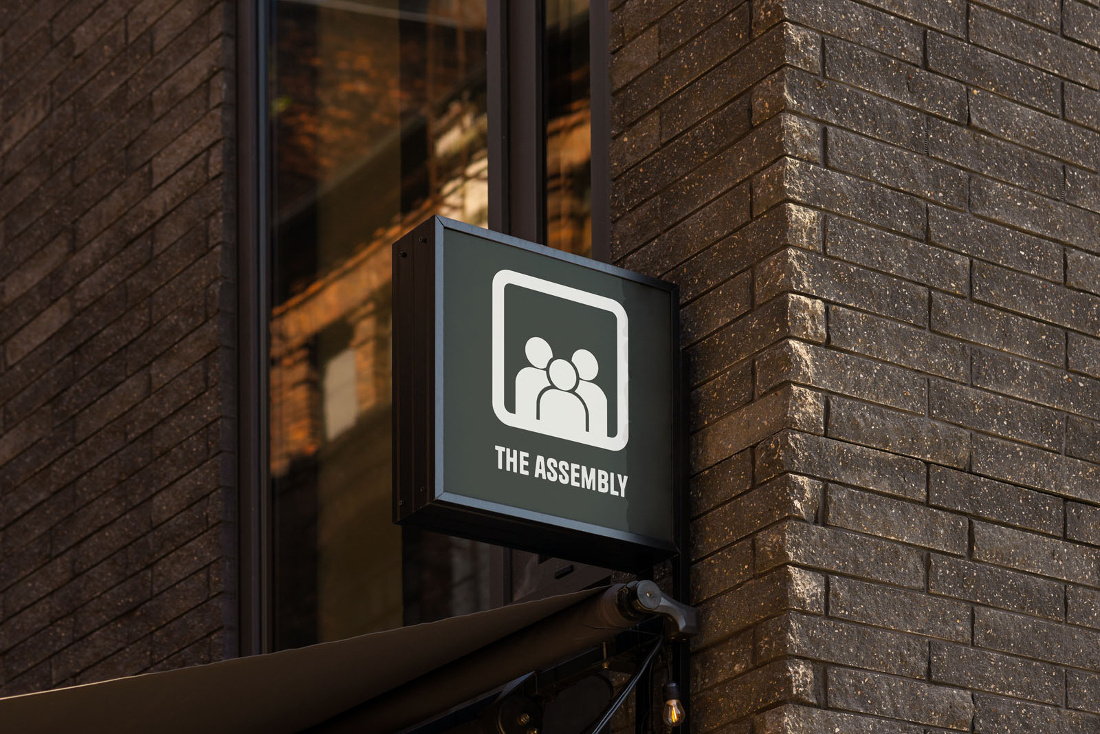 Een zwart vierkant uithangbord aan een donkere bakstenen muur. Op het bord staat een wit icoon van drie personen en de tekst "THE ASSEMBLY", onderdeel van de bewegwijzering van Parc51.