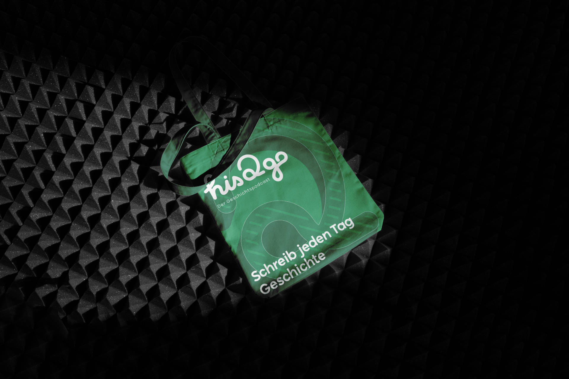 Grüne Stofftasche mit His2go-Logo und dem Slogan „Schreib jeden Tag Geschichte“, fotografiert auf einer schwarzen, texturierten Oberfläche.