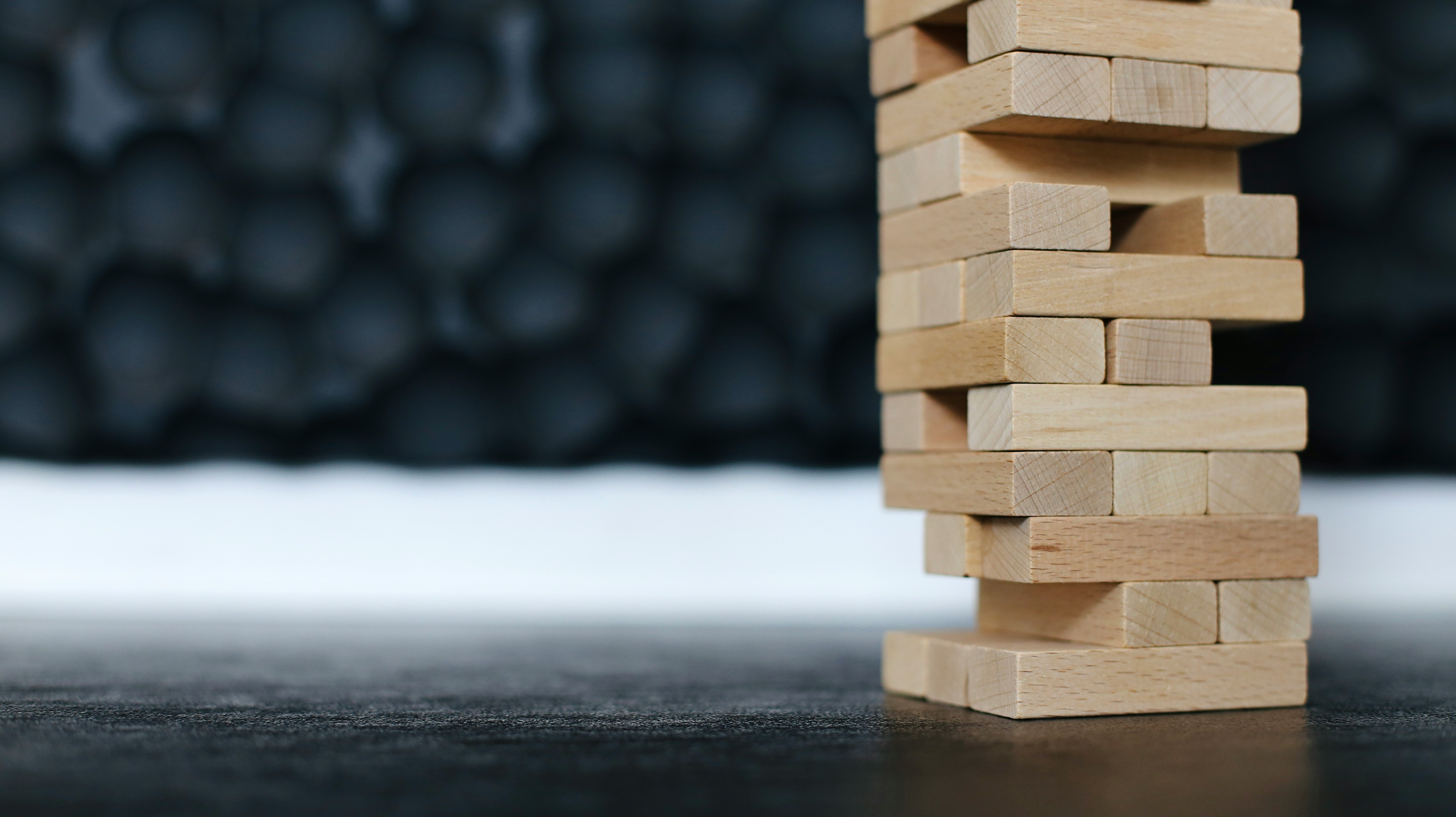 Close-up photo of a wooden Jenga tower game on a dark surface, with blocks carefully stacked and some pieces pulled out, creating a precarious balance