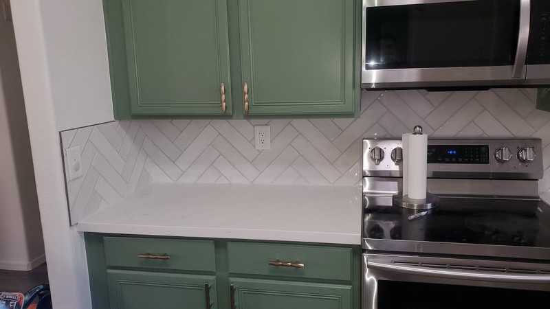 Stylish kitchen with green cabinets, a white herringbone tile backsplash, and stainless steel appliances including an oven and microwave.