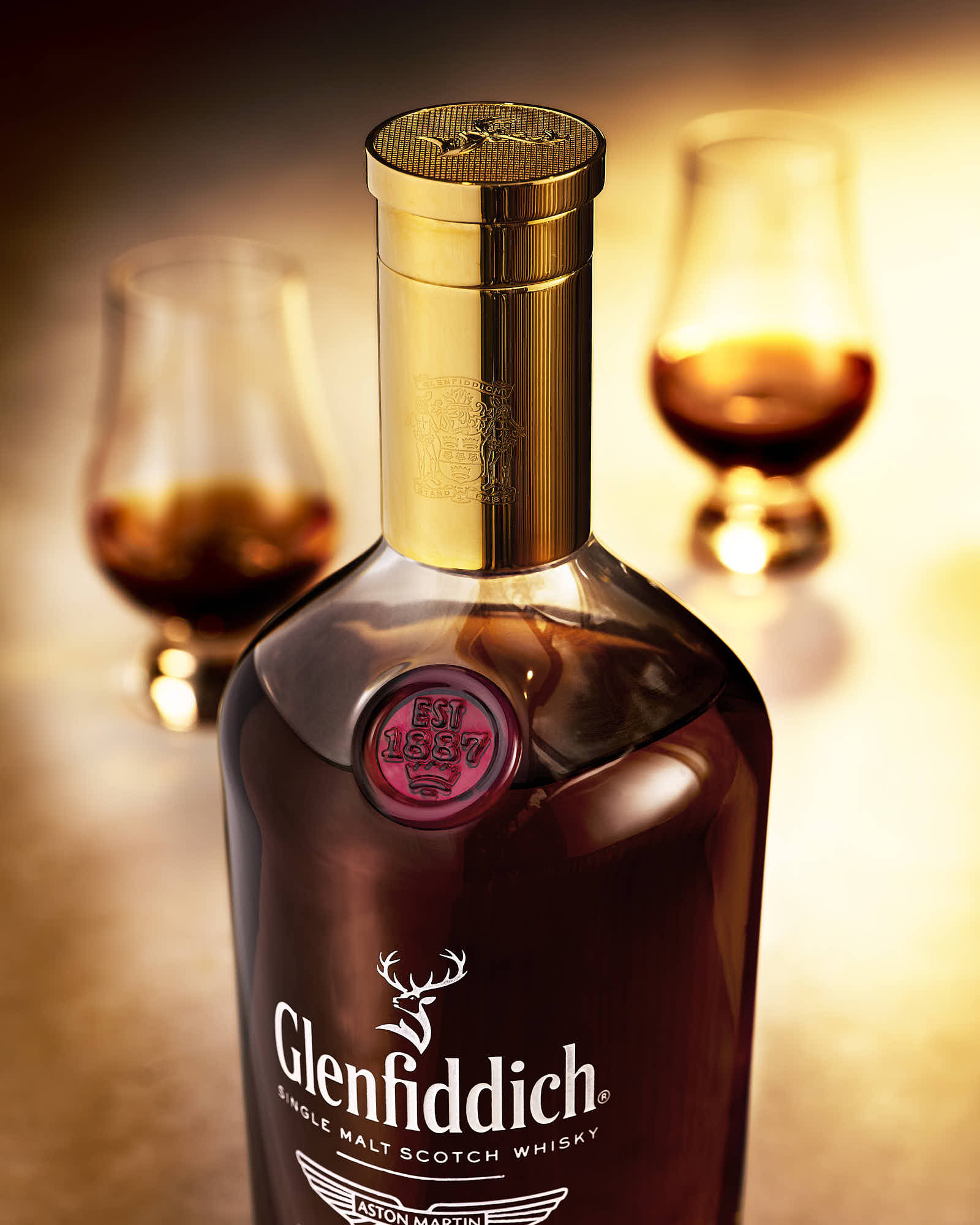 A bottle of whiskey with two glasses in the background, all set against a warm, softly lit background.