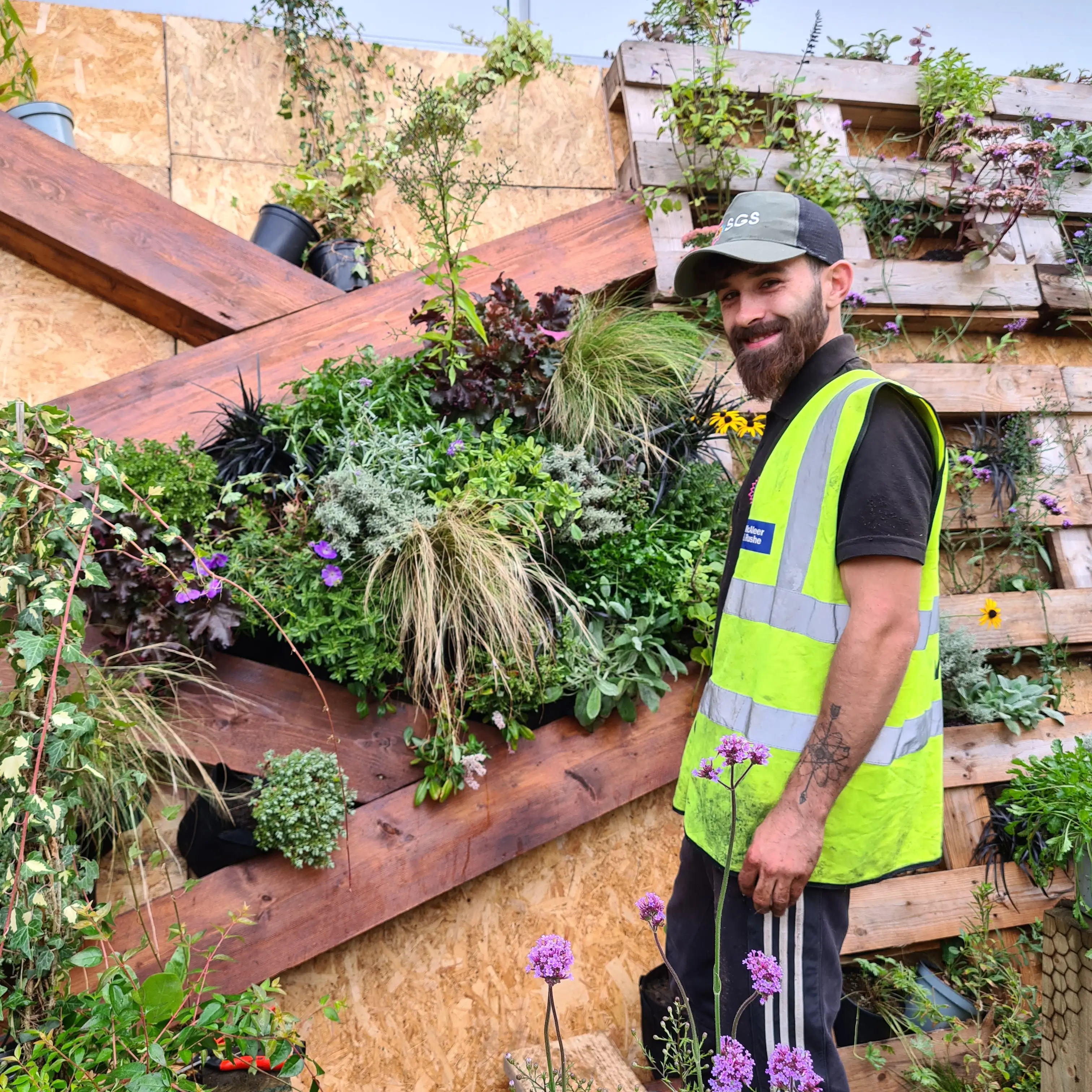 A person in a safety vest stands beside a vertical garden filled with various plants.