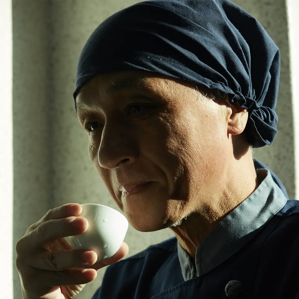 Mistrz Jung Tae-young (정태영) z destylarni Baekkyung, w charakterystycznej niebieskiej czapce, w pełnym skupieniu degustuje trunek z małego, porcelanowego kieliszka