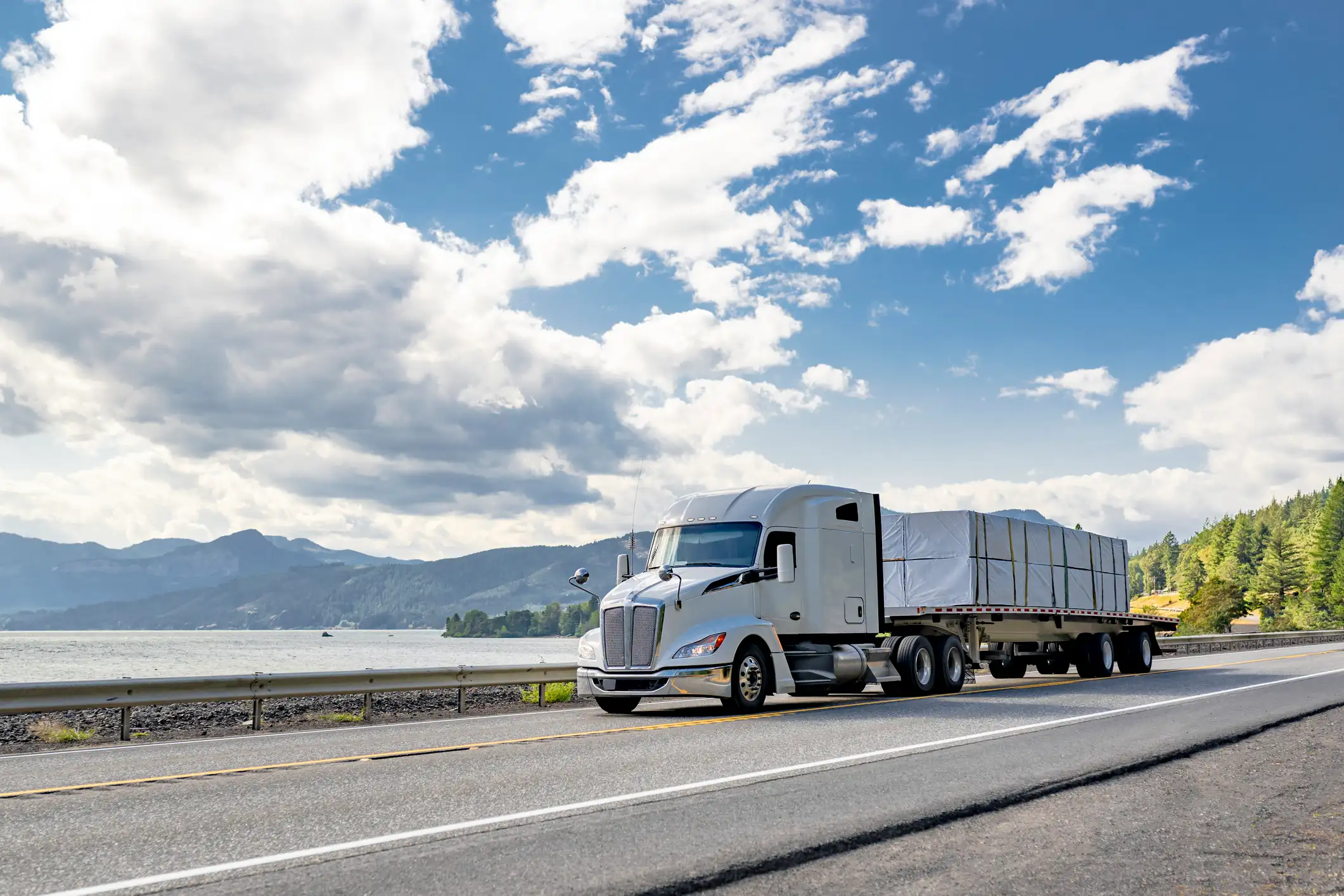 A silver semi-truck hauling cargo along a scenic highway bordered by trees and mountains under a blue sky — representing GIA Group, LLC’s commitment to fast, reliable support for truck insurance needs.