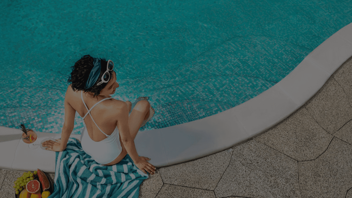 Fotografía de una mujer sentada a la orilla de una piscina, sumergiendo los pies en el agua mientras sostiene una bebida y un plato con fruta.