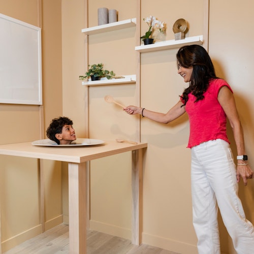 一個男孩的頭從桌子上的一個洞中探出來，而一個女人則在旁邊挑逗性地拿著一把木勺。