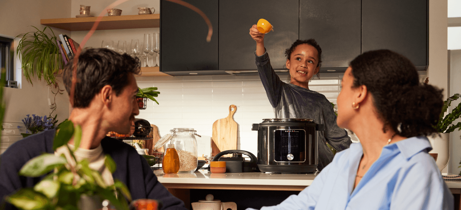 Child lifts a lemon above a multicooker while two adults watch and smile in a modern kitchen
