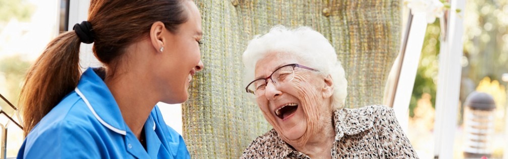 Care worker and elderly woman laughing together in a home setting.