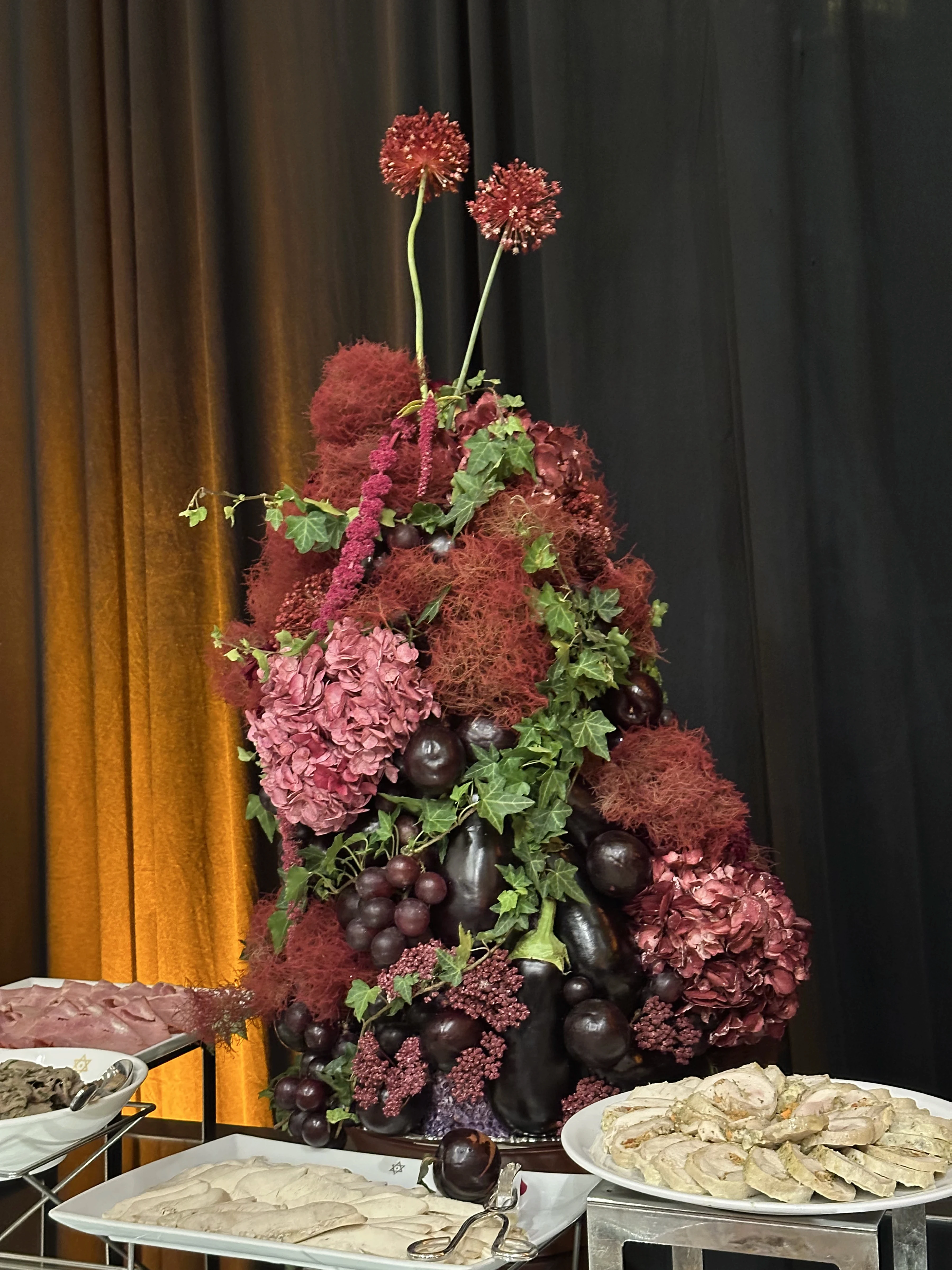 Centro de mesa exuberante con hortensias, uvas, berenjenas y hiedra en buffet con cortinas doradas.