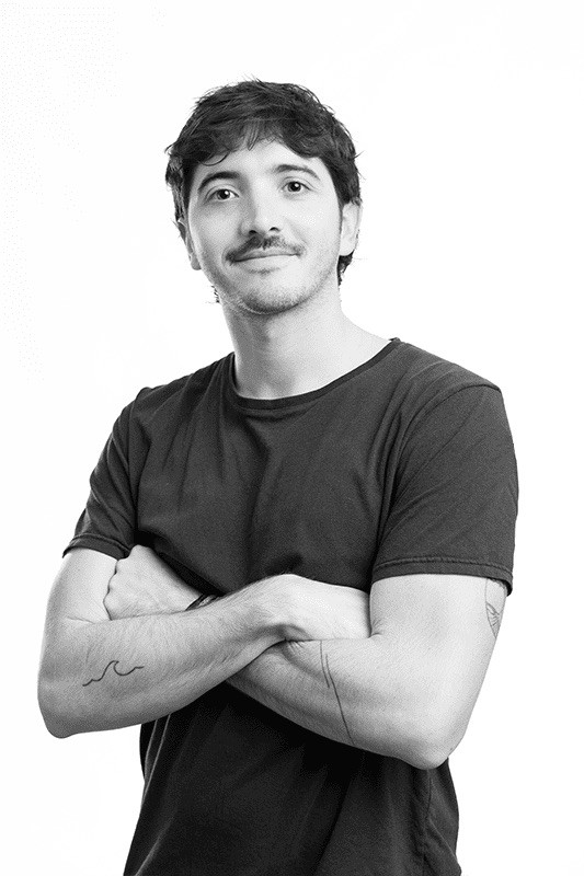 Black-and-white studio portrait of a smiling man with tousled dark hair and a mustache, wearing a black t-shirt with a visible forearm tattoo, arms crossed, against a plain light background.