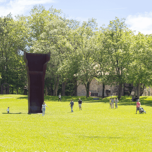 人们在阳光明媚的公园里，绿草和树木环绕的大型户外雕塑附近行走和站立。
