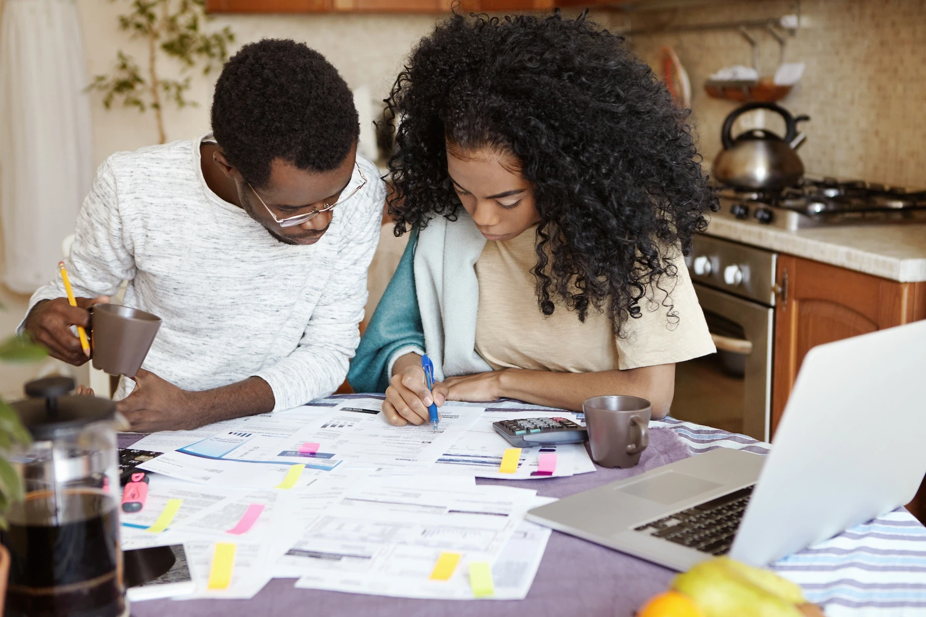 A couple reviewing financial documents together at a kitchen table, surrounded by papers, a laptop, and a calculator.