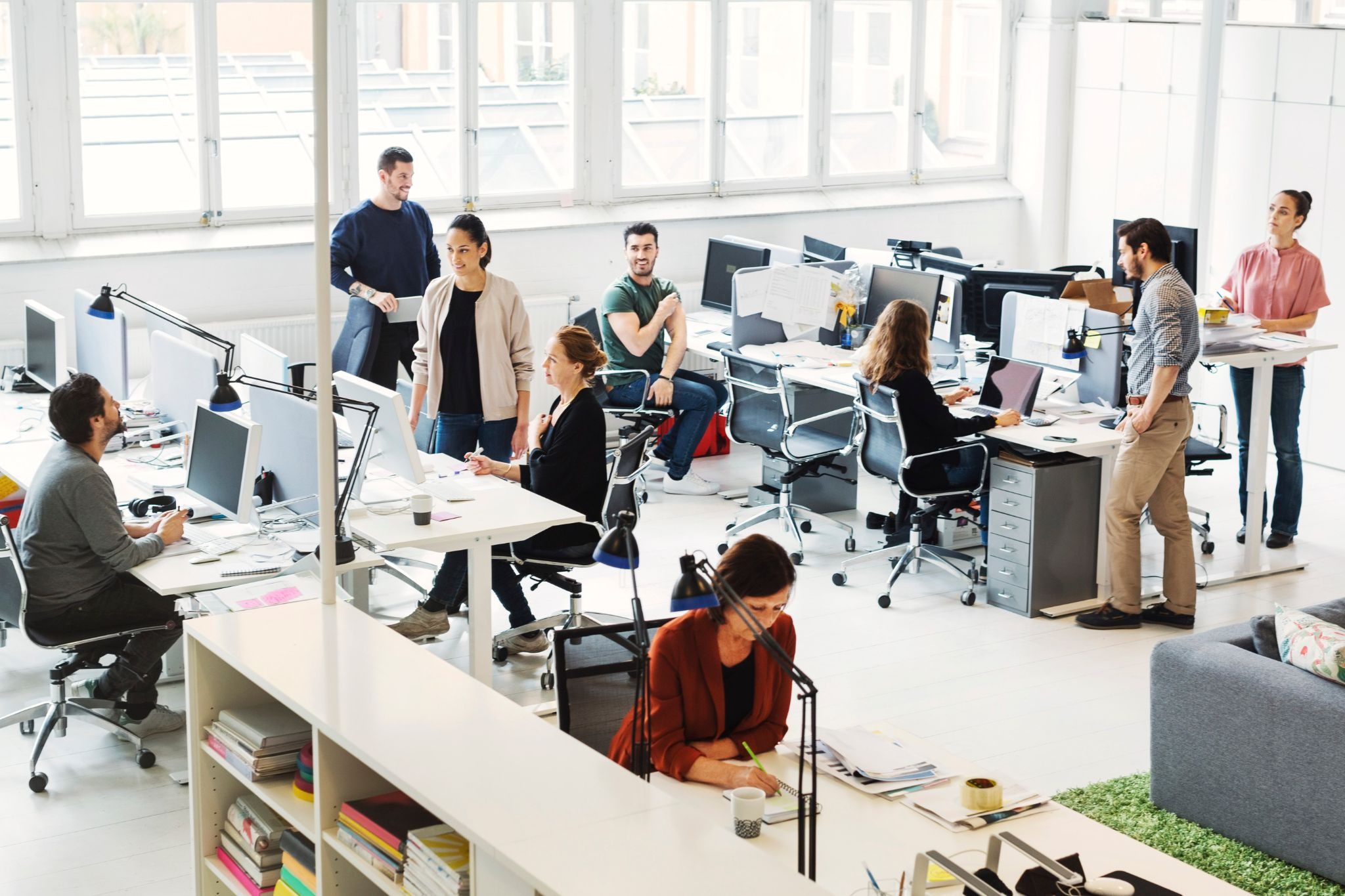 Busy open-plan office with multiple employees working at desks and collaborating in groups.