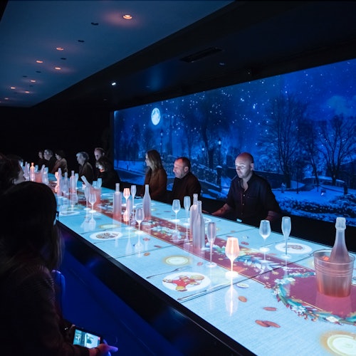 People seated at a long table with illuminated projections of food and drink, under a dark blue night sky scene displayed on the wall.