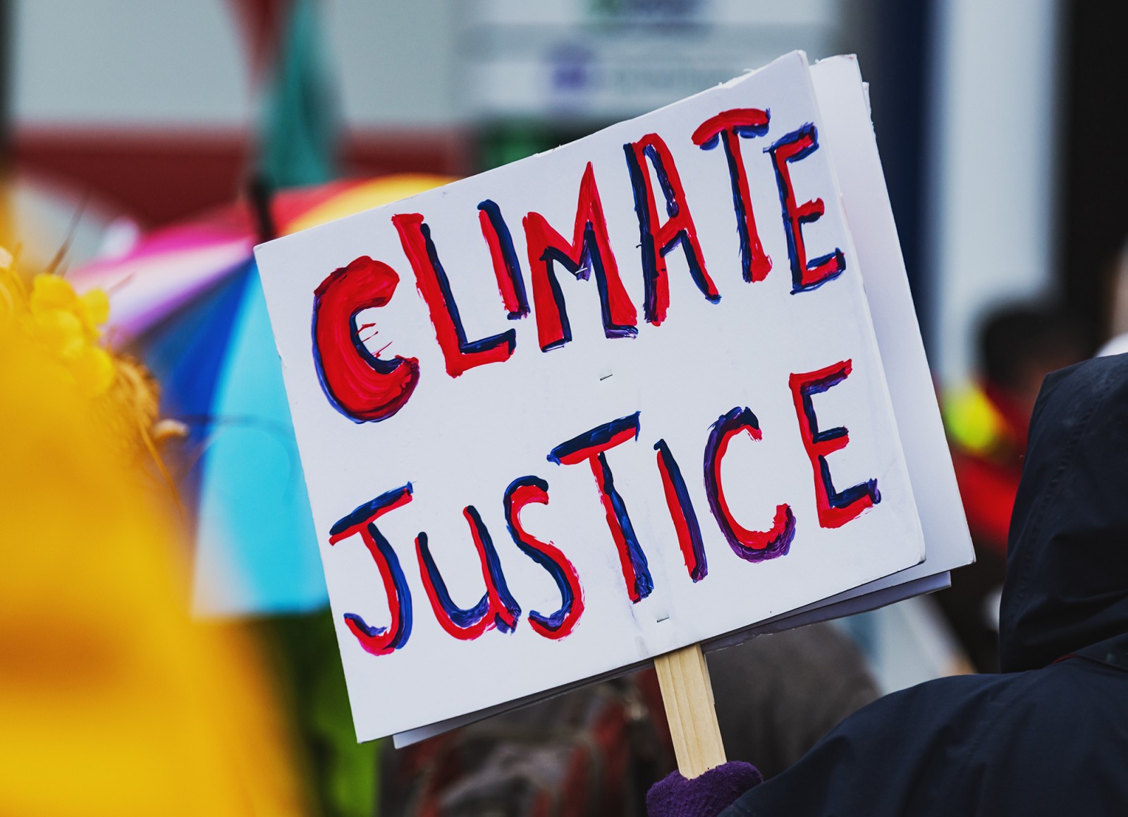 A close-up of a protest sign that reads "CLIMATE JUSTICE," surrounded by people and colorful umbrellas.