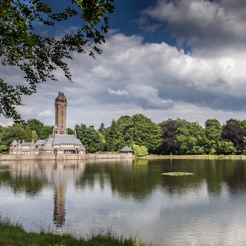 背の高いベージュの塔と建物が、穏やかな湖に映り込み、部分的に曇った空の下で豊かな緑に囲まれています。