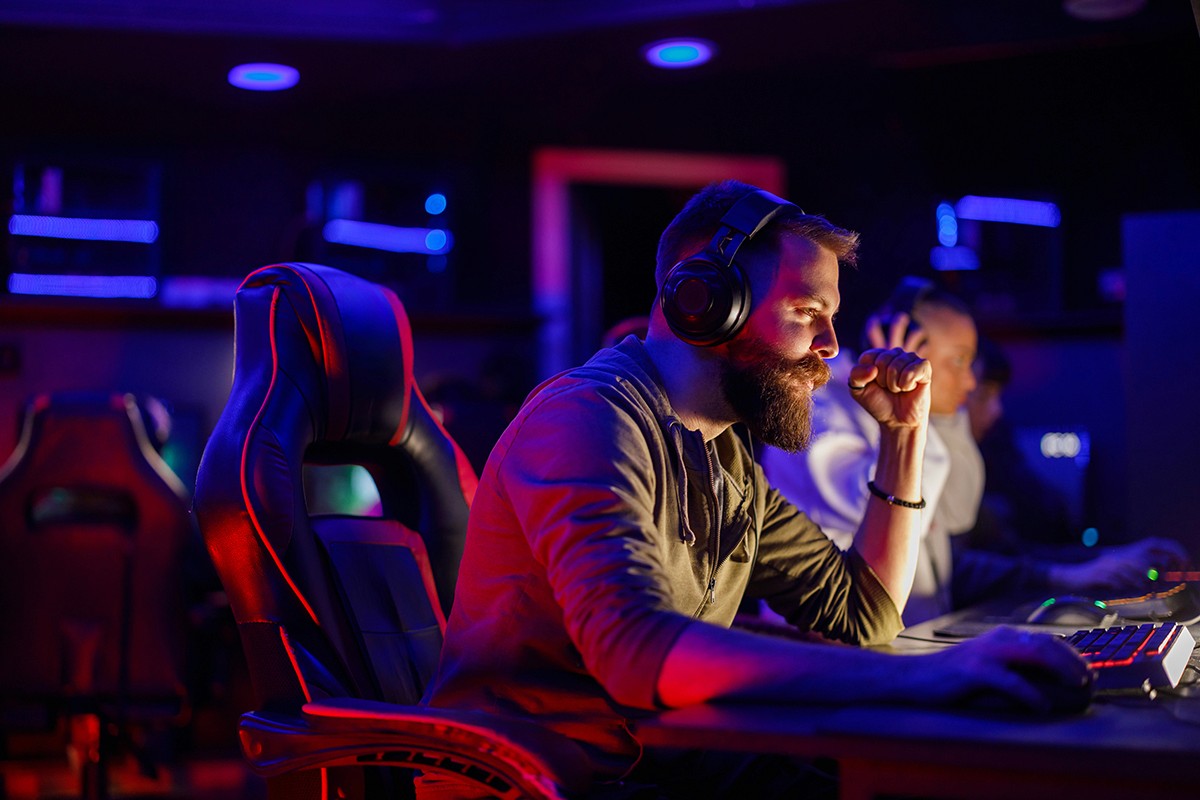 Group of people gathered around a row of computers in a dimly lit, neon-accented space. A person in a white hoodie focuses on the screen, while others stand nearby watching attentively