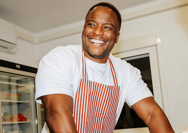 Smiling man wearing a white shirt and red striped apron in a kitchen setting.