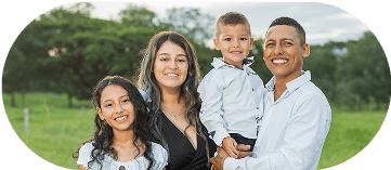 A smiling family of four—a man, a woman, a young boy, and a young girl—posing outdoors in a grassy park at sunset