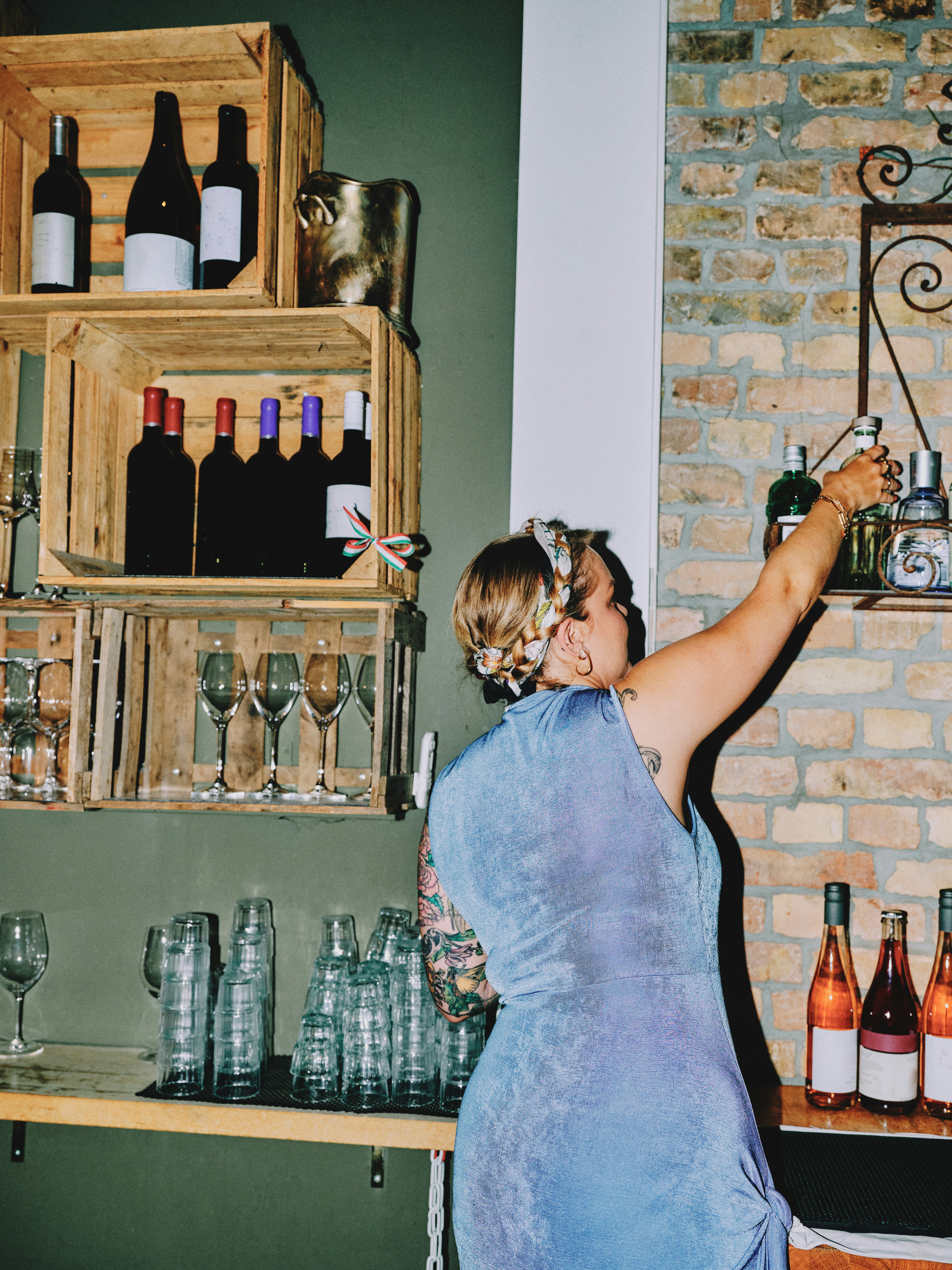 Rear view of a tattooed female bartender in a blue halter dress reaching up to a shelf on an exposed brick wall, wine bottles and glassware surrounding her
