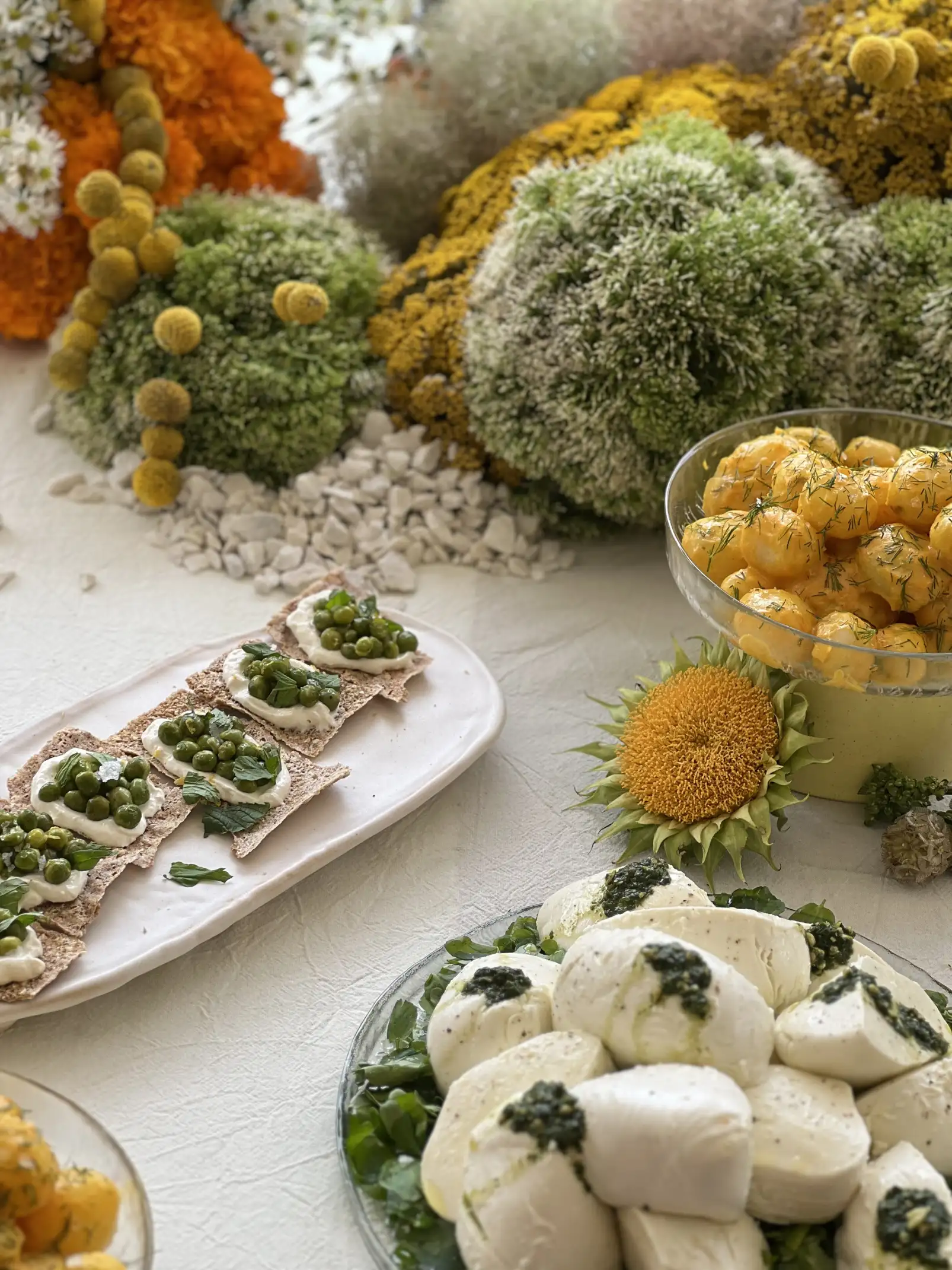 Detalle de mesa con esferas florales verdes, amarillas y naranjas como decoración, acompañada de platos con burrata al pesto, tostadas con crema y arvejas, y un bowl de tomates amarillos.