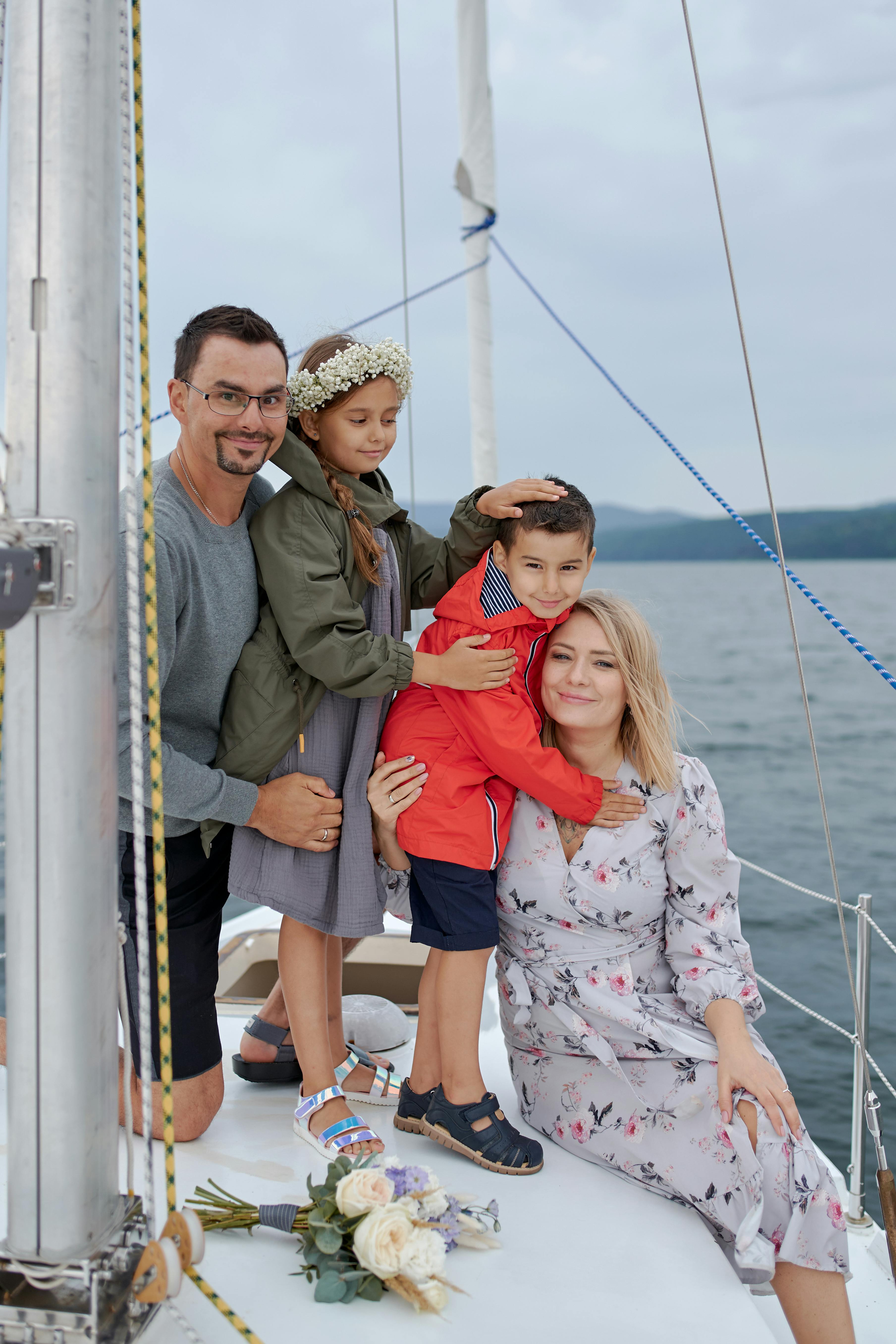 A family poses together on a sailboat with a cloudy sky and calm water in the background, featuring a bouquet of white and pale pink flowers resting on the deck.