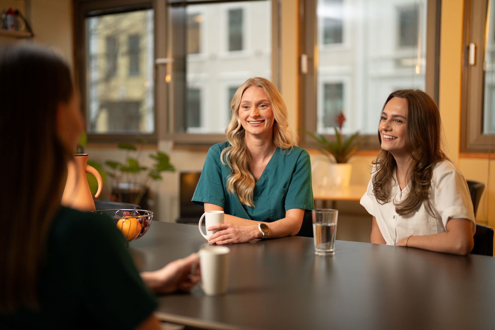 Tre kvinner deltar i samtale ved et bord, med naturlig lys som kommer gjennom vinduene bak dem.