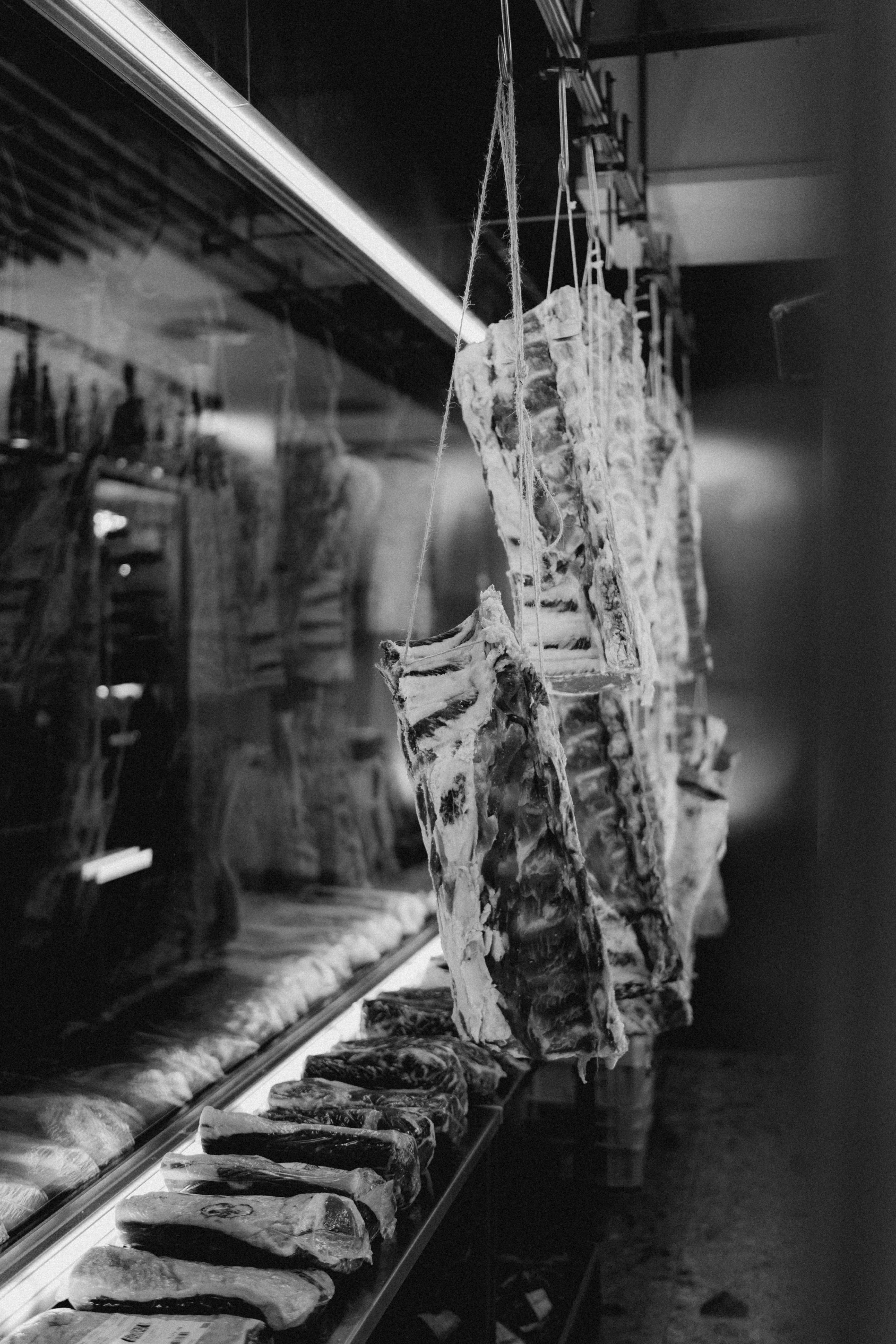 Black and white photography of pieces of meat hanging and lying in a refrigerated room, with the inscription 'MORE MEAT'