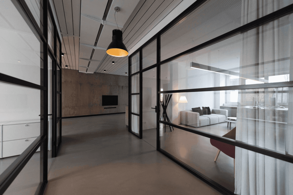 A stylish, modern lounge area featuring pastel green sofas, tan chairs, round wooden tables, and ceiling plants, with large windows and sheer curtains.