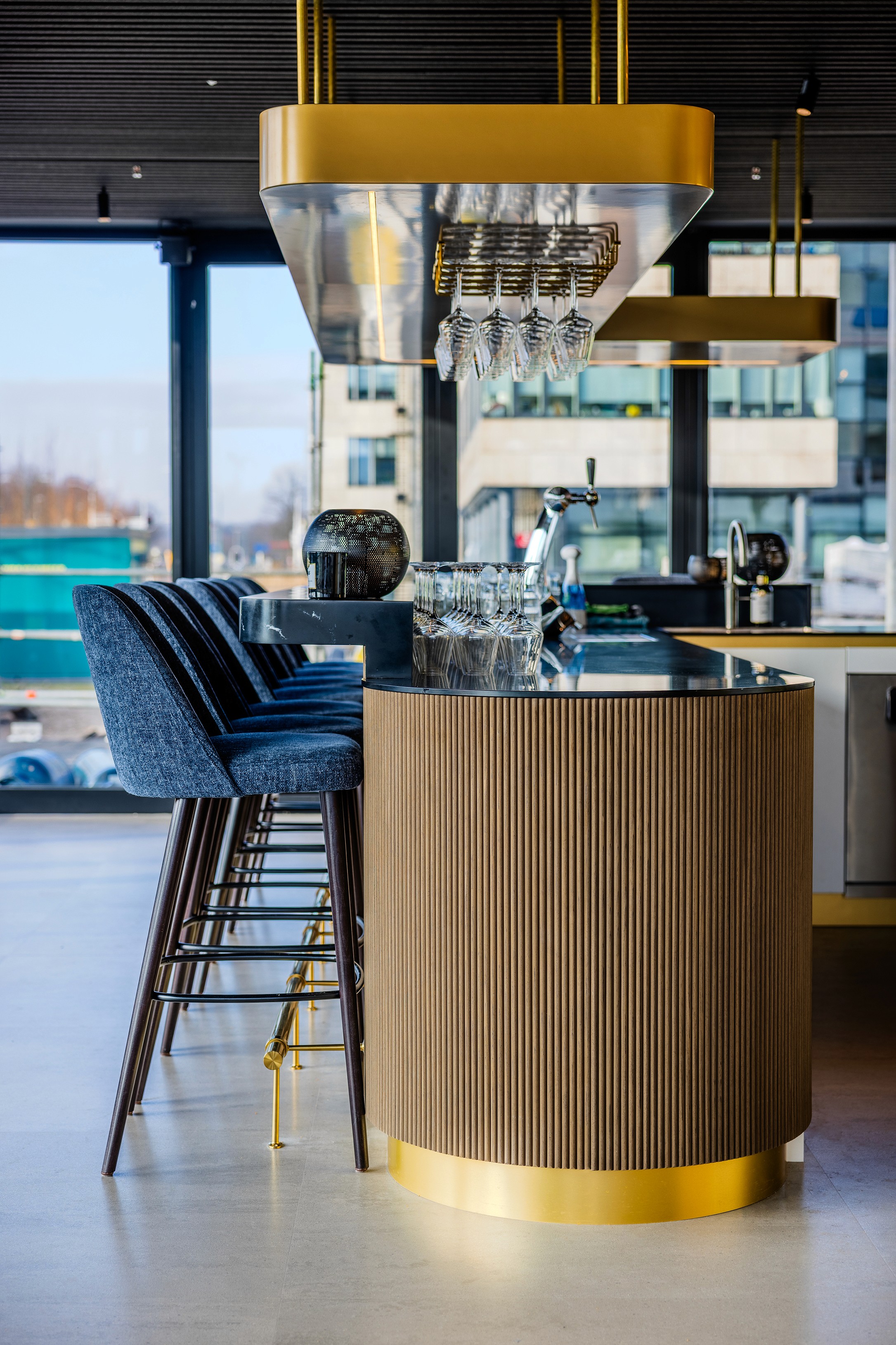 Stijlvolle barhoek in AFAS DotNL met een goudkleurige cilindrische bar, hoge barkrukken en opgehangen wijnglazen tegen een gespiegeld barblad – DSN Workspace Solutions