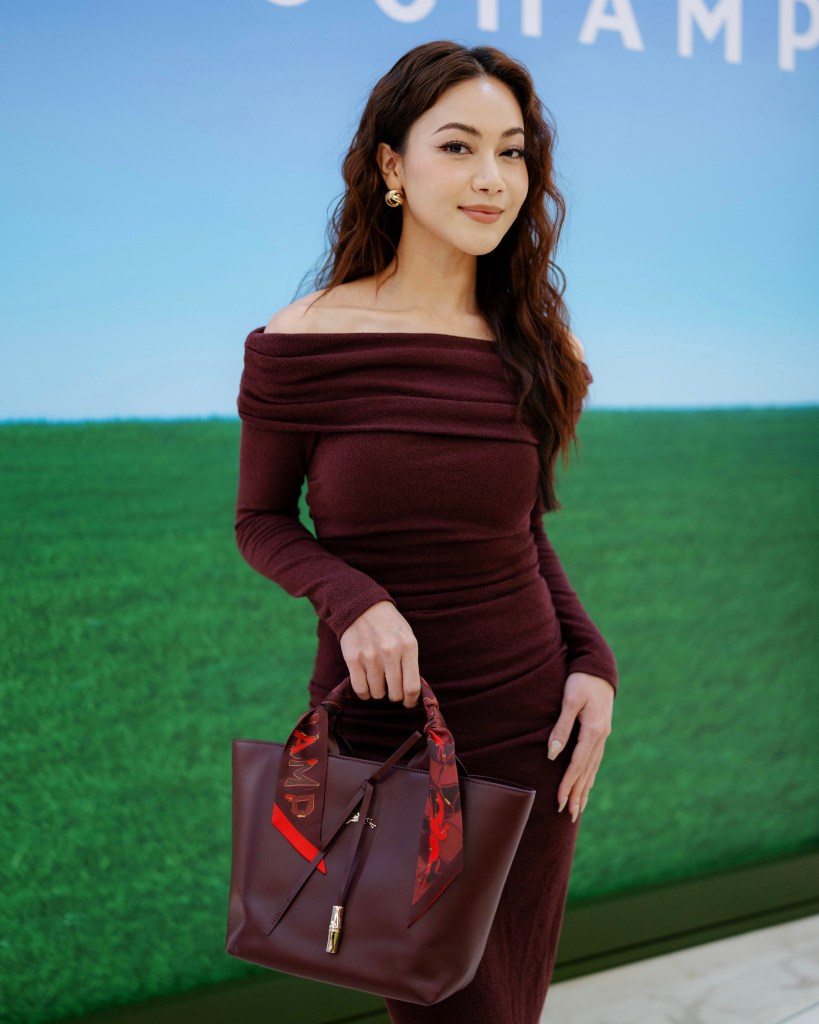 A woman in an off-shoulder burgundy dress holding a brown handbag with a red and black scarf tied to the handle, standing in front of a green and blue background.