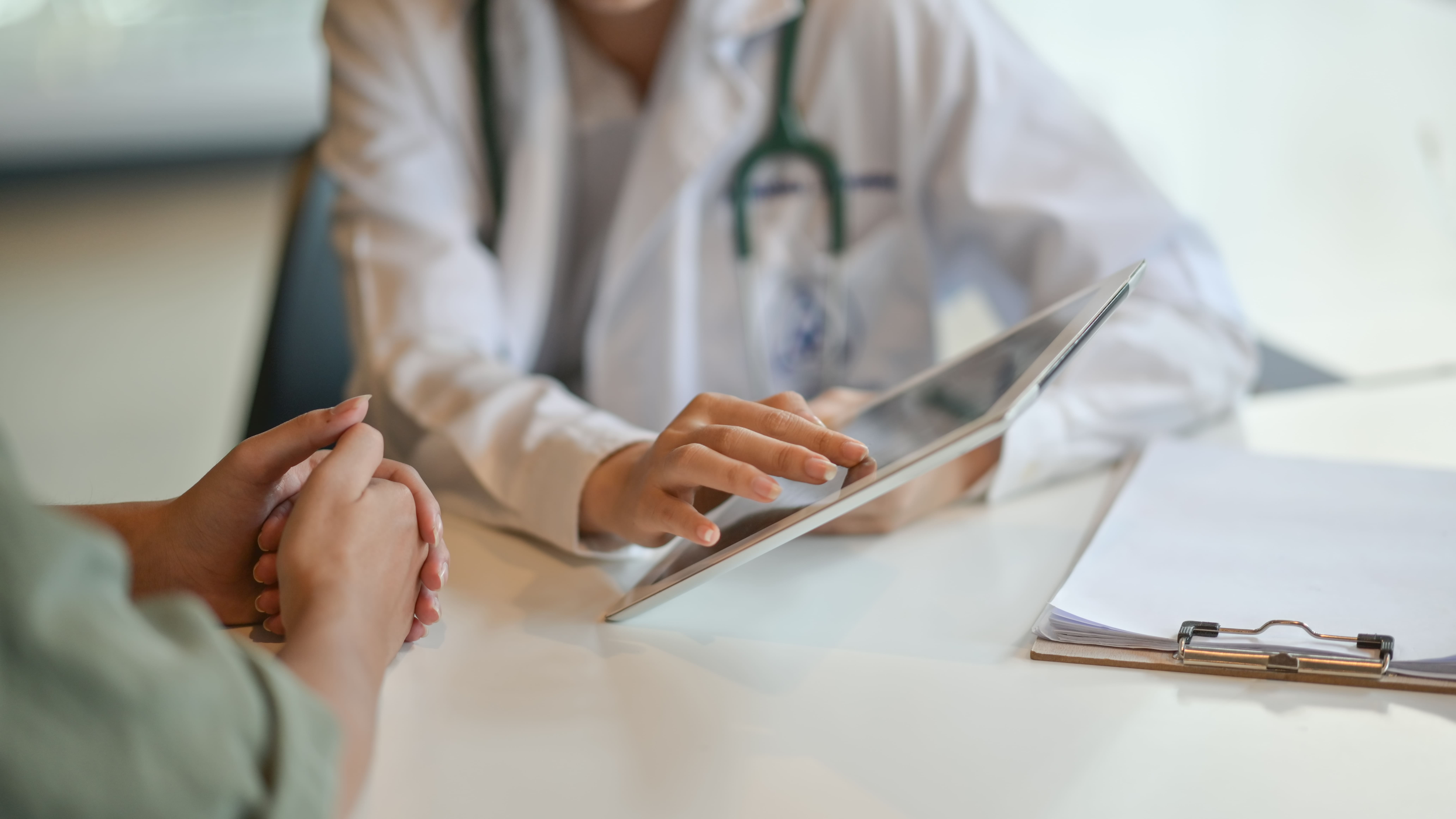 A doctor in a white coat discusses a patient's chart on a tablet across a desk, while the patient listens attentively, highlighting a medical consultation moment.