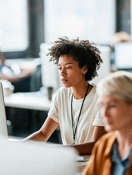 A young person with short, curly hair sits at a desk working on a computer in a bright classroom or office setting. They appear focused on the screen, with other people and desks softly blurred in the background, suggesting a collaborative learning or work environment.
