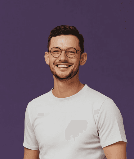 Smiling man with glasses and a short beard wearing a white t-shirt stands against a deep purple background, conveying a cheerful and friendly tone.