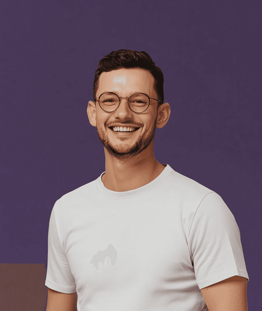 Smiling man with glasses and a short beard wearing a white t-shirt stands against a deep purple background, conveying a cheerful and friendly tone.