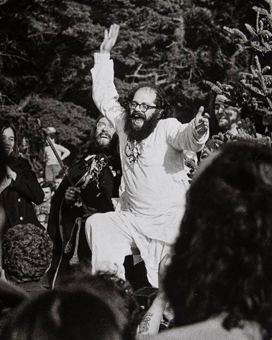 Black-and-white photograph of a bearded performer in white raising his arms before a crowd.