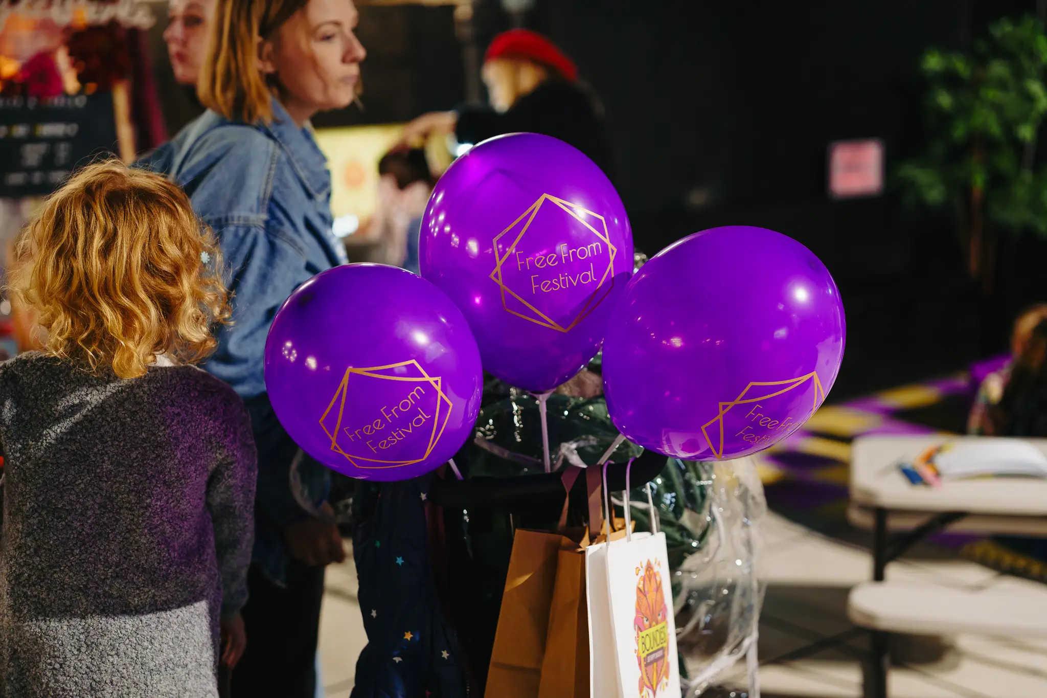 Three purple balloons featuring the gold Free From Festival logo, with attendees walking in the background of the event space.