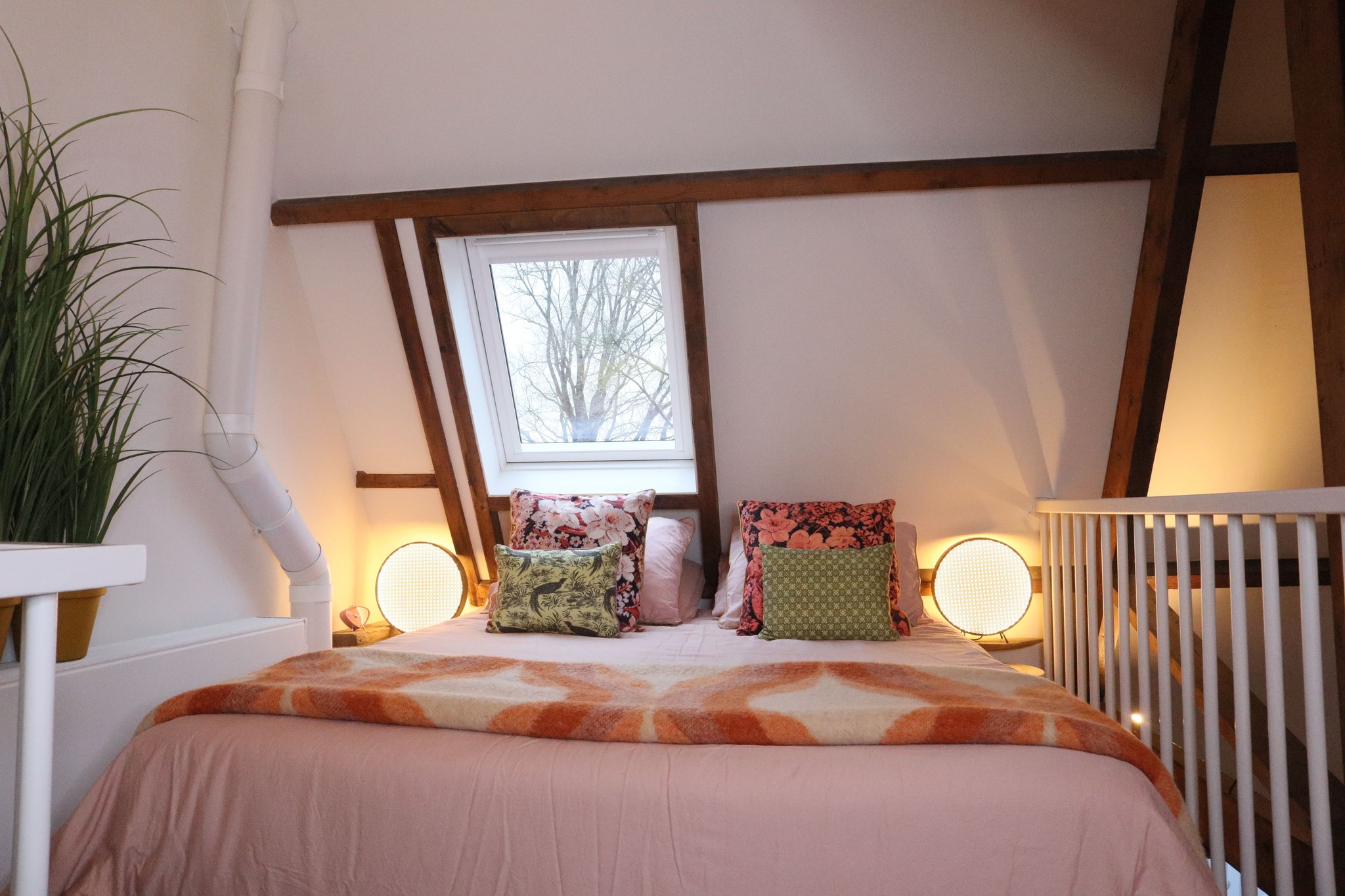 Intieme slaapkamer met een roze beddengoed, kleurrijke kussens en twee ronde lampen naast het bed. Het raam biedt uitzicht op de buitenomgeving en houten balken zijn zichtbaar in het plafond.