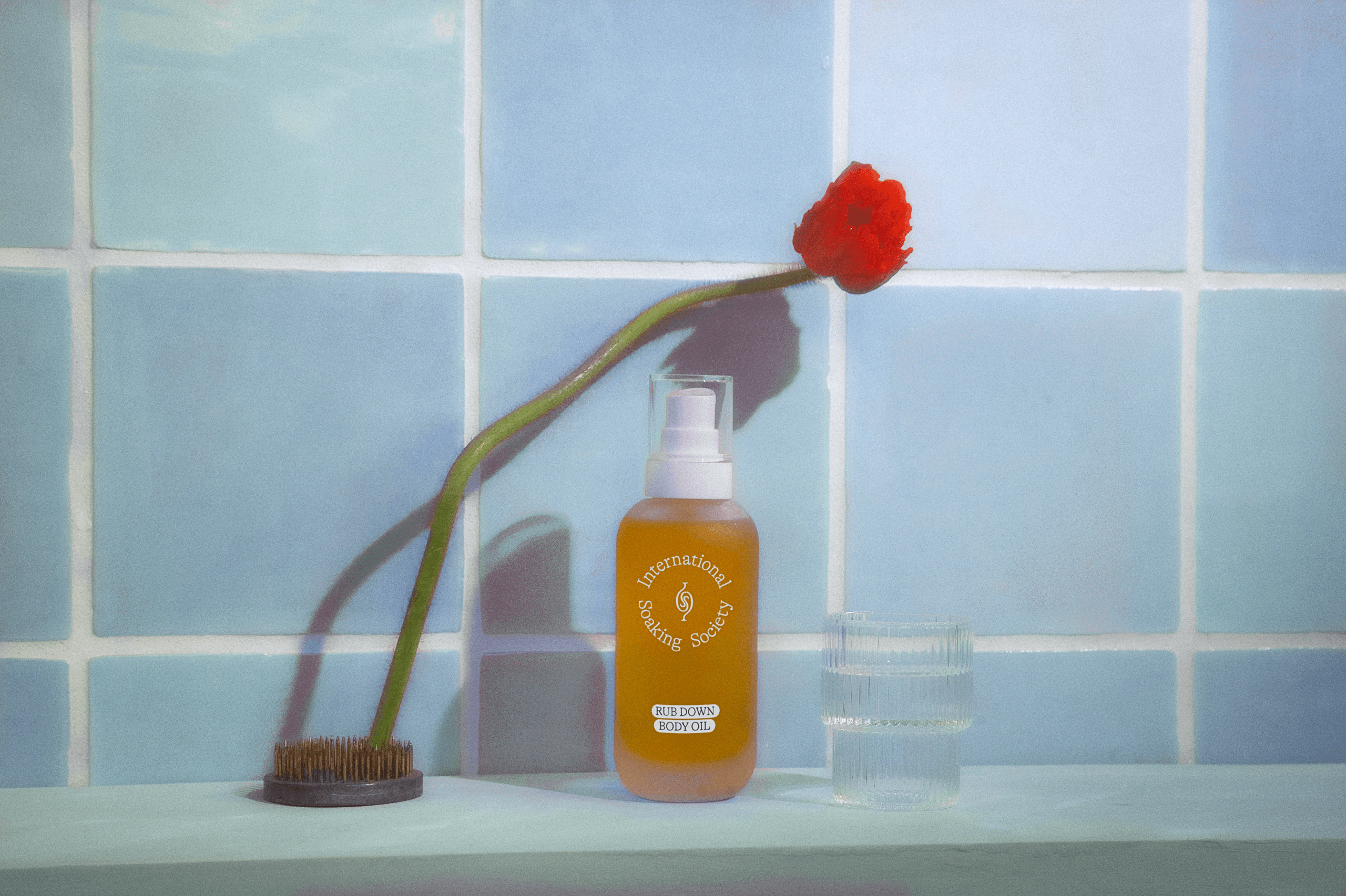 A bottle of International Selling Society's Body Oil sits on a light blue countertop next to a red poppy flower in a dark brown vase. A clear glass with vertical lines is positioned to the right of the bottle. The background is a light blue tiled wall.