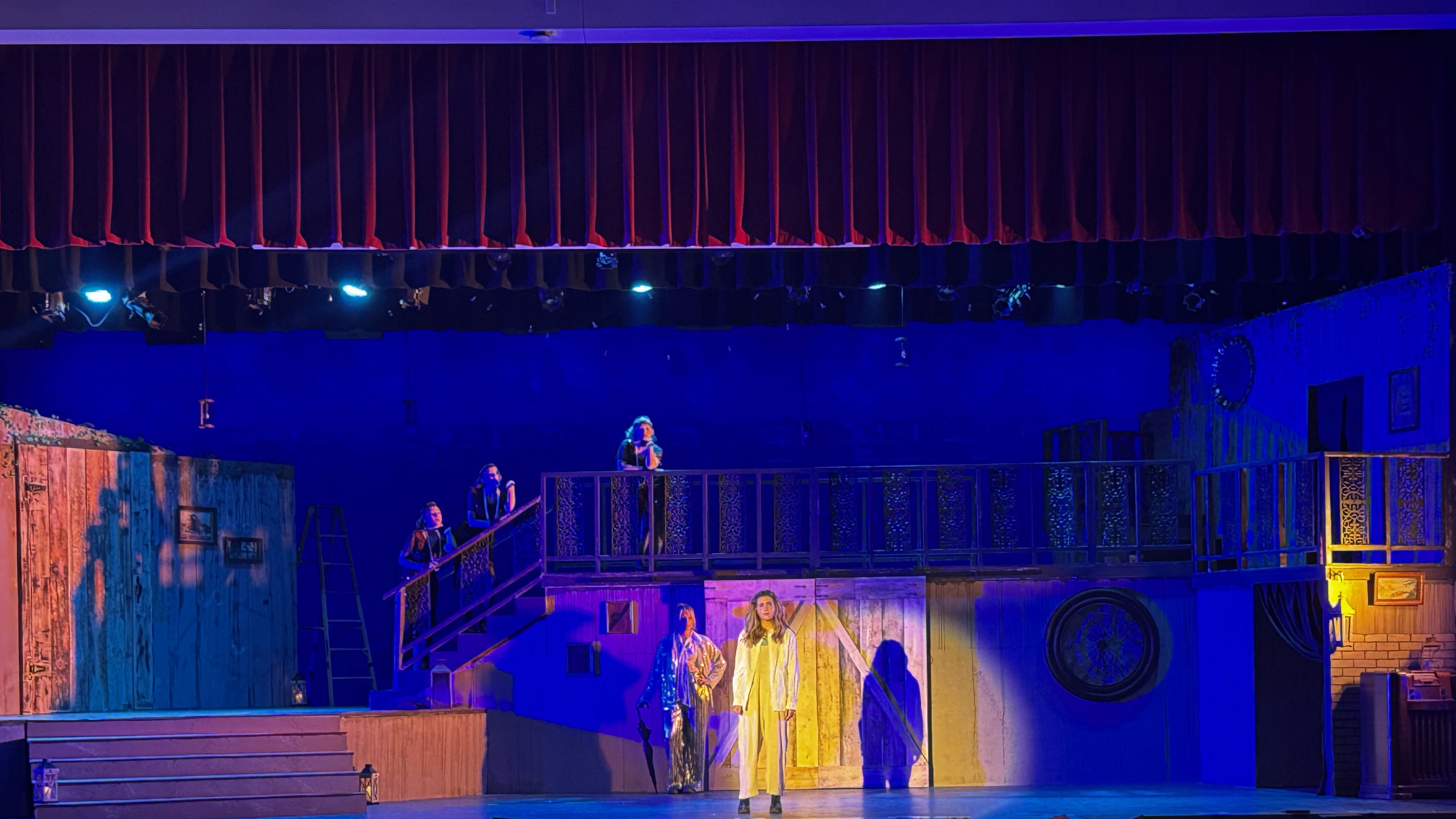 A theater stage is illuminated with dual temperature lighting, featuring a striking blue and amber contrast, highlighting Eurydice, Hermes, and the Fates and enhancing the depth of the set design. The shadows create further blues and purples that pool on the set.