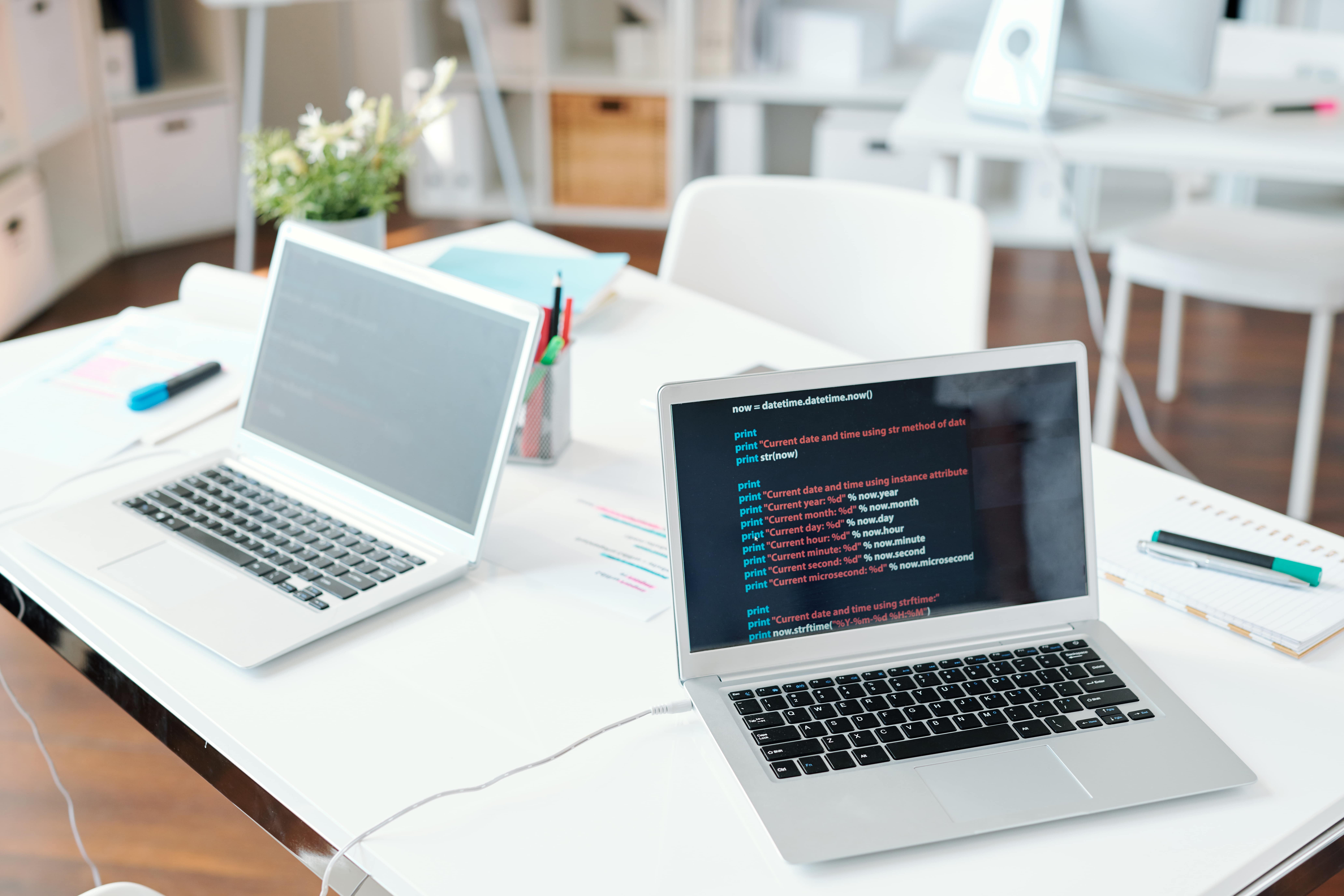 A modern developer's workspace with a silver laptop on a white desk displaying Python code, with a second laptop and office supplies in the background.